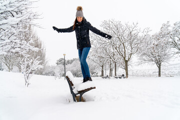 Chica joven en un parque nevado en un día de invierno con gorro de lana gris, jeans y cazadora...