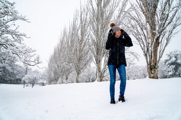 Fototapeta na wymiar Chica joven en un parque nevado en un día de invierno con gorro de lana gris, jeans y cazadora negra