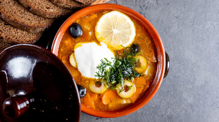 hodgepodge with lemon and sour cream in a clay plate, salt, pepper and rye bread on a gray cement background