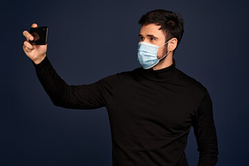 Upset young man in black sweater, with sterile face mask making video call with mobile phone, spreading hand isolated on Pacific Blue background. Coronavirus 2019-ncov sars covid-19 flu virus concept.