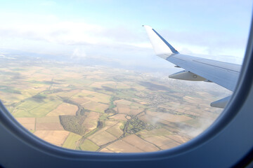 Obraz premium High view from airplane window of landscape of colorful farmland with green, light green and brown soil under white airplane wing, with blue sky white clouds background