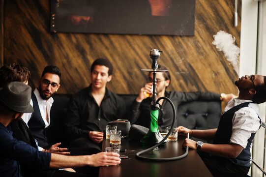 Group of handsome retro well-dressed man gangsters spend time at club, smoking hookah. Multiethnic male bachelor mafia party in restaurant.