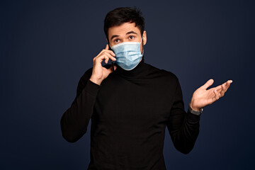 Young man in black sweater, sterile face mask speaking on phone isolated on Pacific Blue wall...