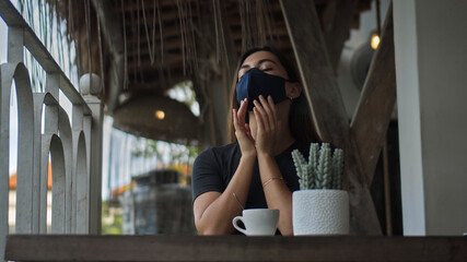 European brunette girl sits at a table in a public place and touches a soft black protective mask with her hands on her face