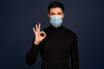 Young man in black sweater, sterile face mask showing OK gesture isolated on Pacific Blue wall...