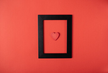 Black photo frame with red wooden heart on the red background. Valentine's Day concept. Flat lay, top view, space for text.