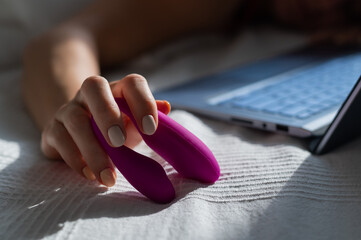 Fototapeta A woman lies in bed holding a clitoral vibrator and watching porn on a laptop. The girl has sex online obraz