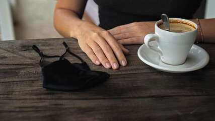 uropean girl sits at a table in a public place with a cup of coffee and nearby to her on a wooden table is a black protective mask