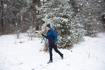 Skier with a backpack and hat with pompom with ski poles in his hands on background of a snowy forest. Cross-country skiing in winter forest, outdoor sports, healthy lifestyle, winter sports tourism.