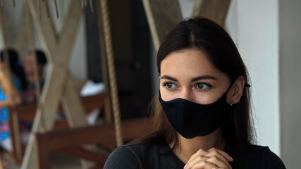 European brunette girl sits at a table in a public place on a black protective mask on her face