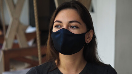 sad European brunette girl sits at a table in a public place on a black protective mask on her face