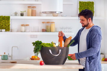 happy young adult man unpacking the shopping bag full of fresh, tasty food at the sunlit home kitchen