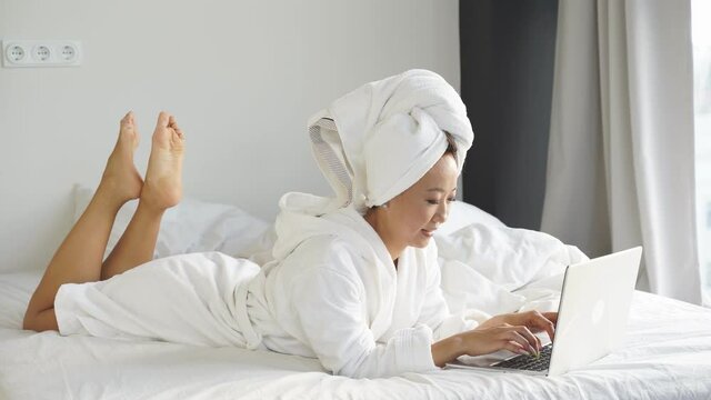 beautiful young Chinese woman working with a laptop, lying in bed at home, working remotely online due to the coronavirus pandemic.