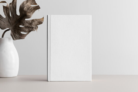 White book mockup with a monstera leaf in a vase on a beige table.