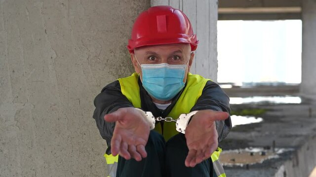 Slow Motion Handcuffed worker in red hard hat and medical protective mask sits on concrete floor of construction site. He looks, talks and gesticulates at the camera