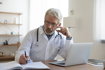 Focused serious middle aged old male physician working with medical prescriptions and laptop, busy mature doctor gp managing patient's appointments or writing illness history at hospital office.
