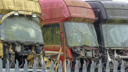Line of scrapped transport vehicles of different colours stored together. All lorries in distressed, weathered state with broken and dismantled elements. Stacked in open air behind metal railing. UK. - 404326950