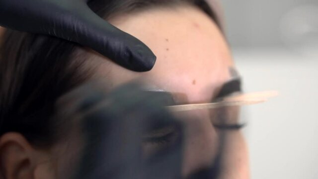 Correction of a shape of eyebrows with hot wax. Brow master applying wax on the eyebrow of female face of a brunette woman. Wax correction of the shape of the eyebrows. Beauty industry