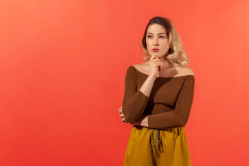 Portrait of a young woman in brown blouse pondering with a serious expression. She touching her chin and thinking with thoughful face, looking away. Indoor studio shot isolated on red background