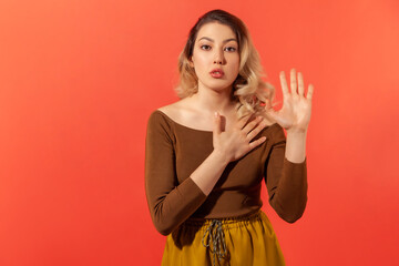 Portrait of serious honest young woman with blond curly hair raising one arm and putting on chest another making oath, swearing. Indoor studio shot isolated on red background