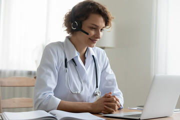 Pleasant happy young female doctor physician wearing headset with microphone, looking at laptop...