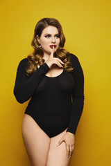 Plus size sexy model girl, with bright makeup, in black bodysuit saying hush and posing at the yellow background. Young plus size woman in black bodysuits isolated on the yellow background.