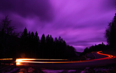 Fototapeta na wymiar Car light lines on a mountain asphalt road in the evening, trees, cloudy sky. Jeseniky mountains,Czech Republic. .