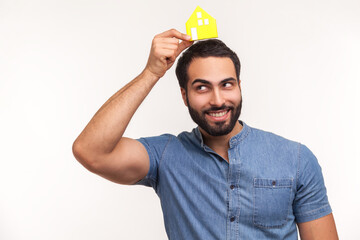 Naklejka premium Happy dreamy man with beard holding little paper house on head, imagining his own apartment, housing purchase, real estate. Indoor studio shot isolated on white background