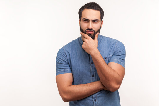 Thoughtful bearded man touching his chin, making difficult decision, facing with dilemma and trying to make choice, awkward situation. Indoor studio shot isolated on white background