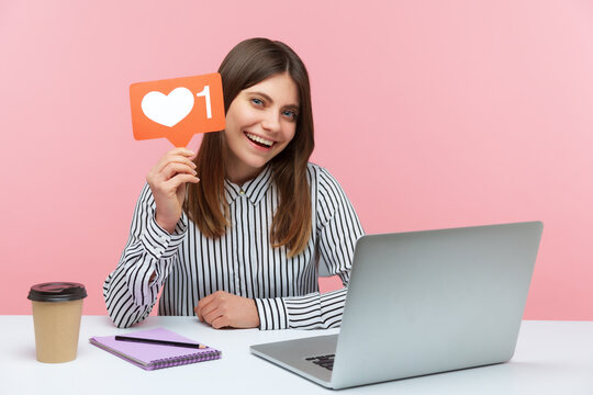 Positive smiling woman blogger holding like counter template sitting at workplace with laptop, making post in social network and waiting for feedback. Indoor studio shot isolated on pink background