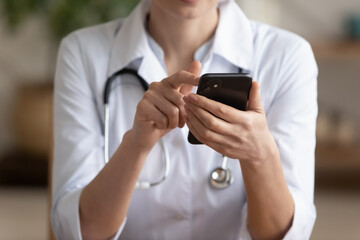 Close up young female medical worker in white uniform holding smartphone in hands, involved in online communication with patient, using mobile applications, modern technology and medicine concept.