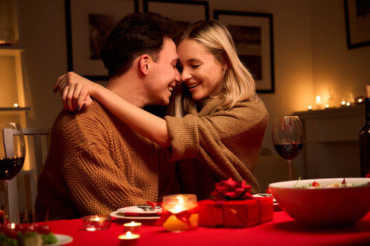 Happy Smiling Affectionate Young Couple In Love Hugging Laughing, Drinking Wine, Talking, Celebrating Valentines Day Dining Together, Having Romantic Dinner Date In Candle Light Sitting At Home Table.