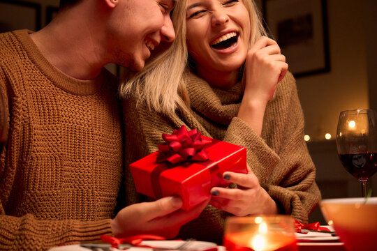 Happy young woman receiving gift box from loving man. Young couple in love hugging, laughing, celebrating Valentines day dining enjoying romantic dinner date holding present surprise sit at home table