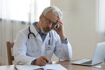 Stressed middle aged old family doctor general practitioner in white uniform and eyeglasses working...