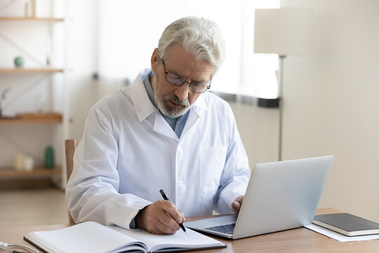 Focused Middle Aged Male Doctor In White Uniform And Eyeglasses Leaving Notes In Paper Journal, Managing Patient's Checkup Meetings Or Prescribing Illnesses Treatment, Using Computer At Clinic Office.