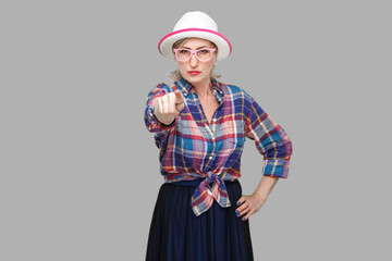 Hey you. Portrait of serious bossy modern stylish mature woman in casual style with hat and eyeglasses standing, pointing and looking at camera. indoor studio shot isolated on gray background.