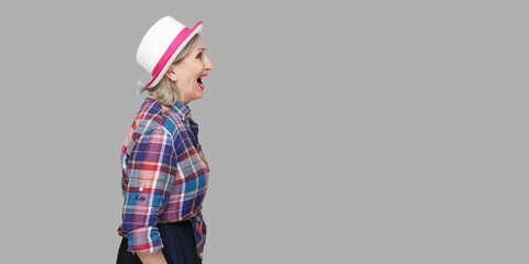 Profile side view portrait of angry modern stylish mature woman in casual style with hat standing, looking forward and screaming. indoor studio shot isolated on gray background.