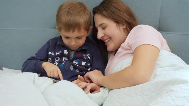 Portrait of happy smiling mother with little toddler son playing on tablet computer while lying in bed