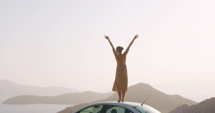 Young woman staying on car roof on road side on top of mountain and raises arms into air, happy, youth and happiness in fornt of sea landscape. Free as a bird concept. Road trip adventure