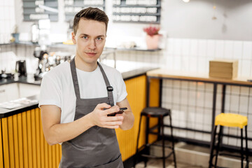 Fototapeta na wymiar he holds the phone in his hands. the employee of the staff of restaurant cafe bartender with dark hair. a man of Caucasian appearance in a work apron and a white T-shirt. posing at work.