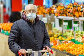 Old senior european man wearing protective facial mask pushing shopping cart in the supermarket. Shopping during COVID-19 concept.