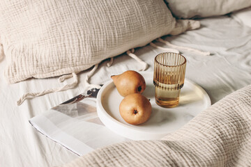Autumn, summer breakfast in bed composition. Glass of water, folded newspapers and pear fruit on white marble tray. Champagne beige muslin cotton bed linen. Velvet cushions. Lifestyle, interior.