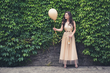 Teen girl in glamorous golden dress standing by the green wall holding balloon.