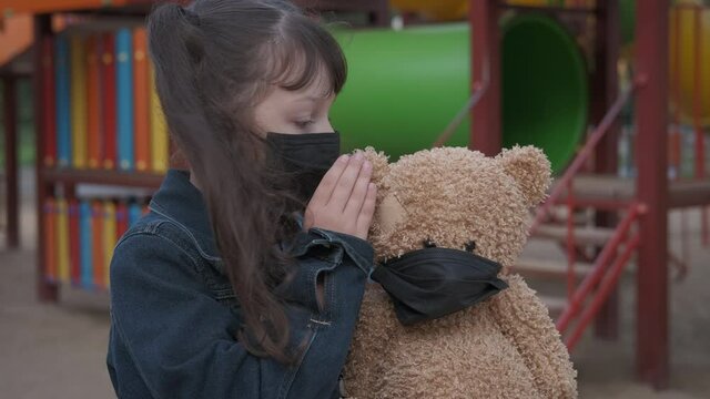 Child talk with teddy. A little happy child in mask speak on teddy bear ear on playground. A concept of isolation and loneliness during quarantine.