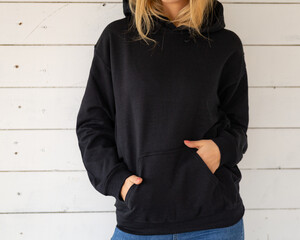 Sweatshirt hoodie mockup. Unrecognizable woman poses in a black sweatshirt against the background of white boards, facing the camera. - 404345911
