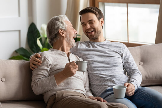 Overjoyed mature father and adult son sit relax on couch at home drink coffee or tea talking on weekend. Happy elderly dad and grownup man child have fun laugh enjoying family time together.
