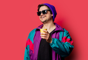 Funny guy in 80s style jacket and sunglasses holds paper cup on red backgorund