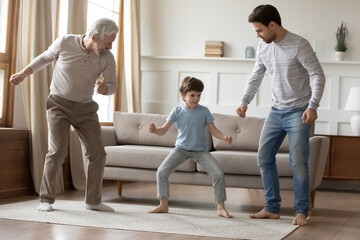 Overjoyed three generations of men have fun dancing together in cozy living room. Happy little boy with young father and elderly grandfather enjoy good weekend day at home. Family concept.