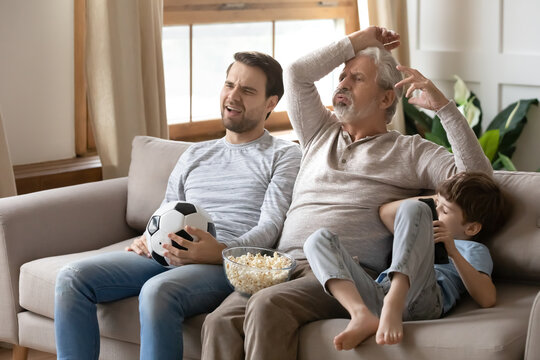 Upset Three Generations Of Men Stressed With Favorite Team Losing Watching Football Match On TV Together. Unhappy Sad Small Boy With Father And Grandfather Feel Distressed With Game Result.