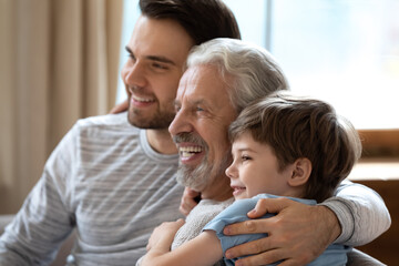 Close up of smiling boy, young father and elderly grandfather relax together in cozy home on weekend. Happy three generations of men look in distance dreaming of happy future. Family concept.